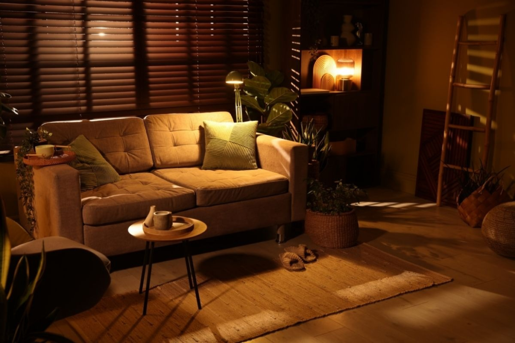 Warmly lit living room featuring a tan sofa, green pillows, and indoor plants.

