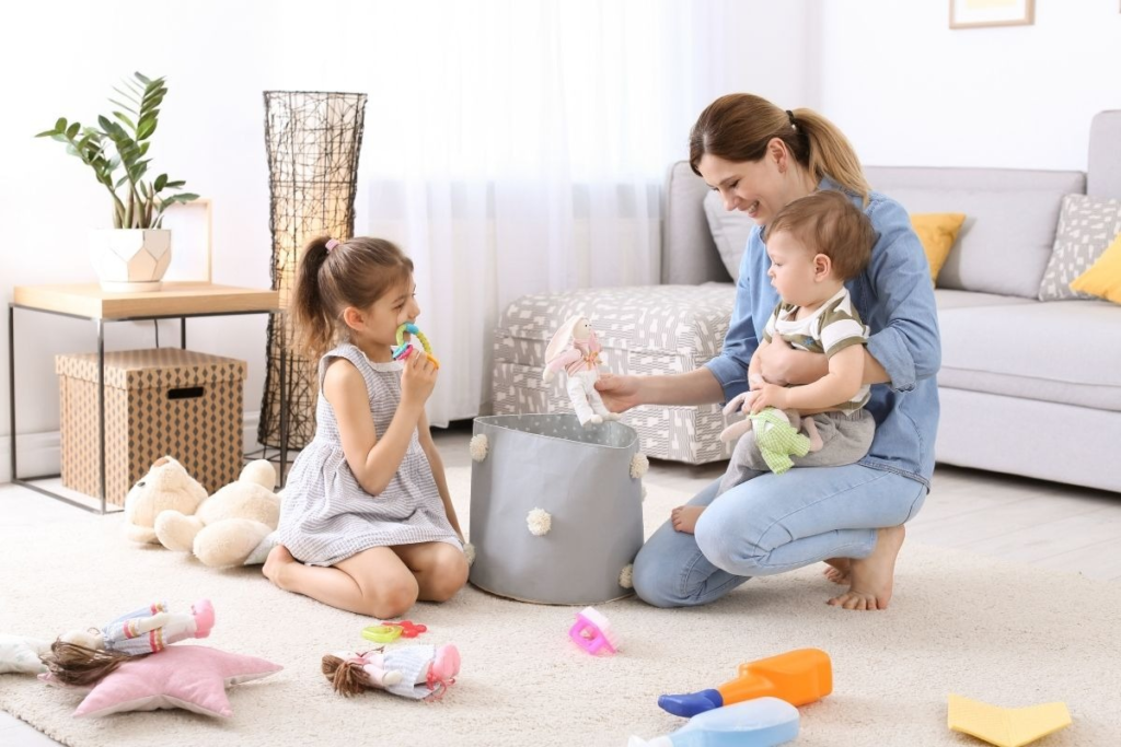 A woman kneels on the floor holding a baby while a child sits nearby, surrounded by toys and a storage bin. 