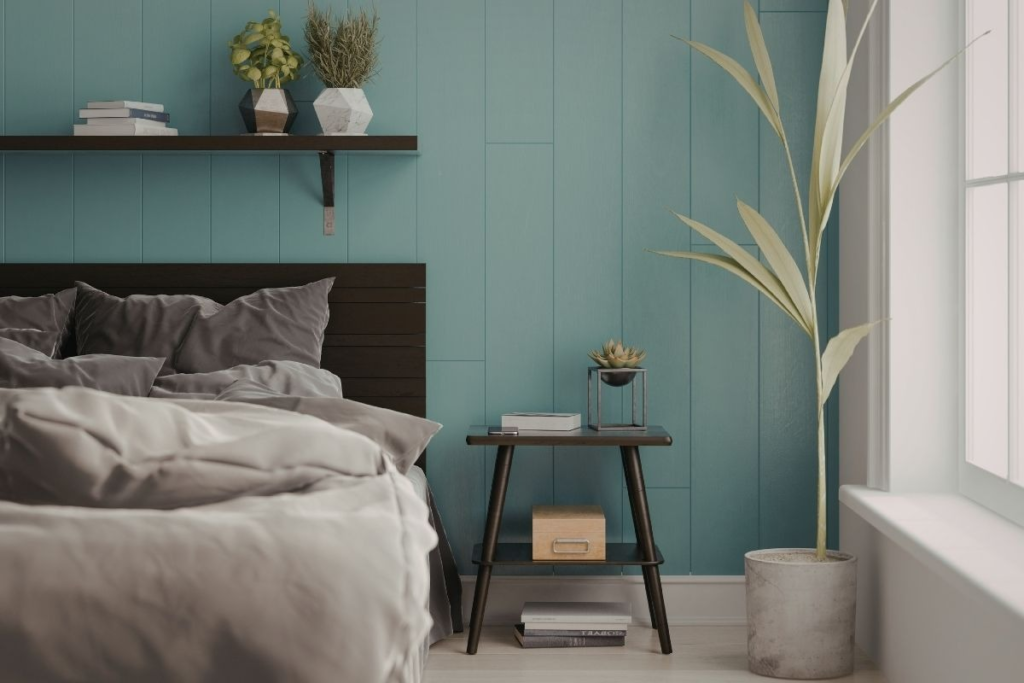 A bedroom with a made bed, a dark wood headboard, and a teal paneled wall. A floating shelf above the bed holds books and two potted plants. A small side table next to the bed has a plant and a box on it, with books below. A large potted plant stands by the window.