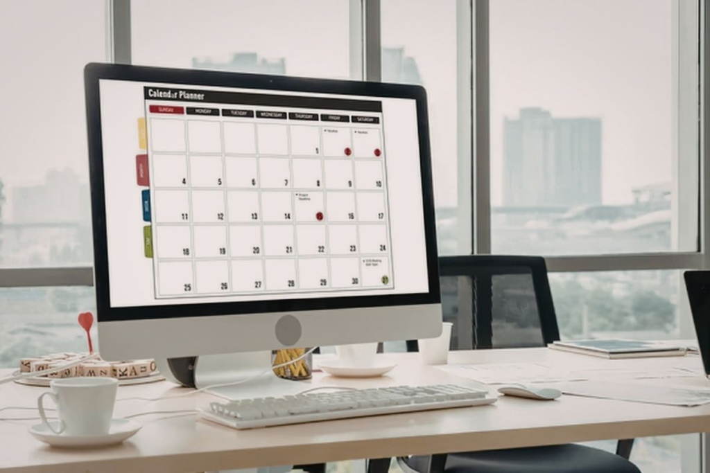 A desktop computer displays a digital calendar planner with marked dates, placed on a desk in an office setting by a window.