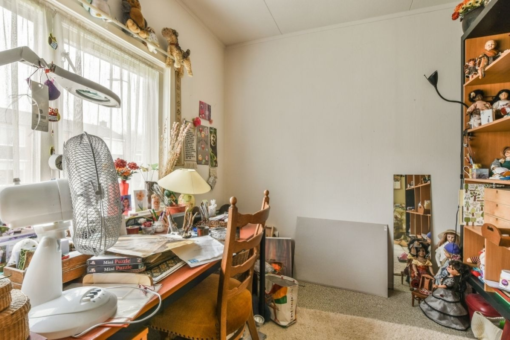 A cluttered desk with a fan, lamp, papers, and books in front of a window, with shelves full of dolls and stuffed animals in the background.