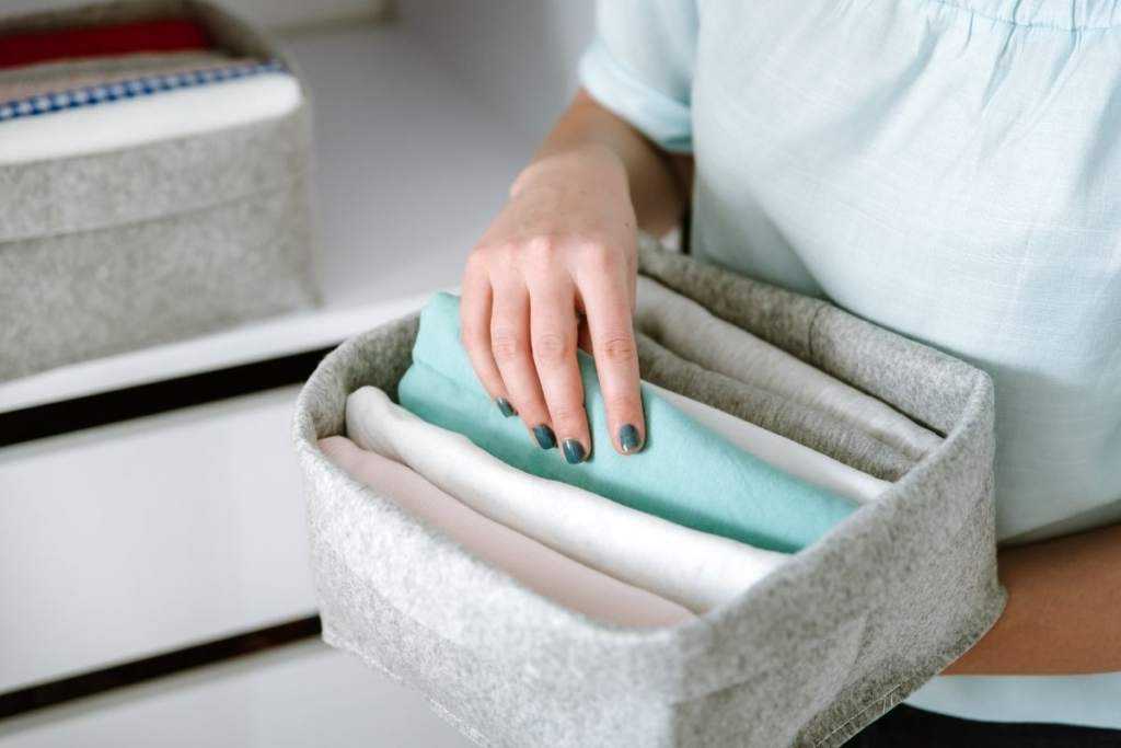 A hand places a folded teal fabric into a gray fabric storage bin filled with folded white and pink fabrics.