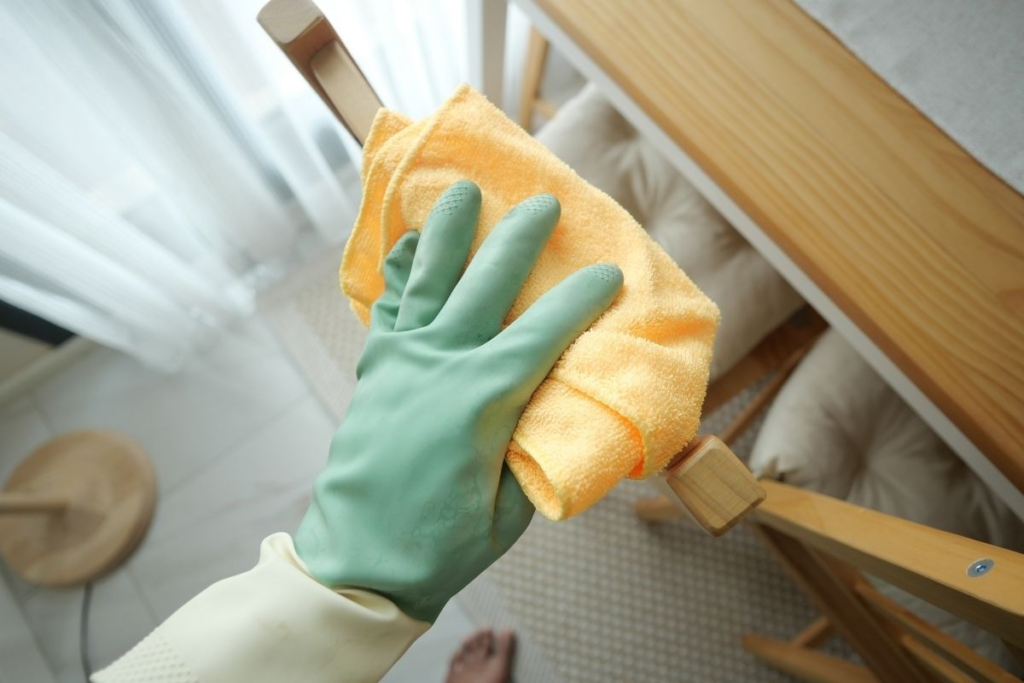 A hand wearing a green cleaning glove wipes down a wooden chair frame using a yellow microfiber cloth