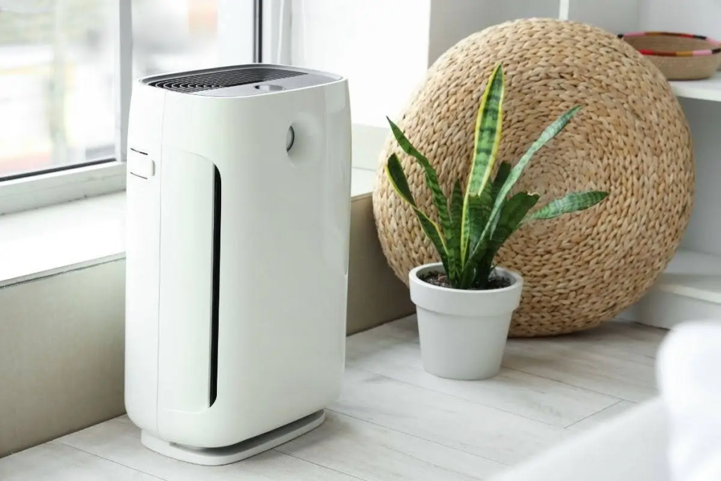 White modern air purifier sitting on a light wooden floor near a potted snake plant and a woven pouf.