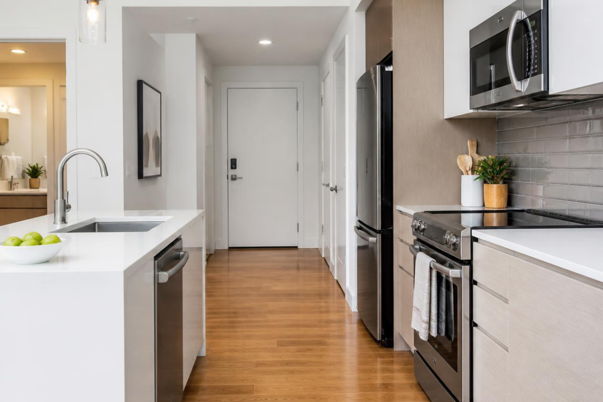 Modern apartment kitchen with stainless steel appliances, white countertops, and warm hardwood flooring leading down an entryway hallway.