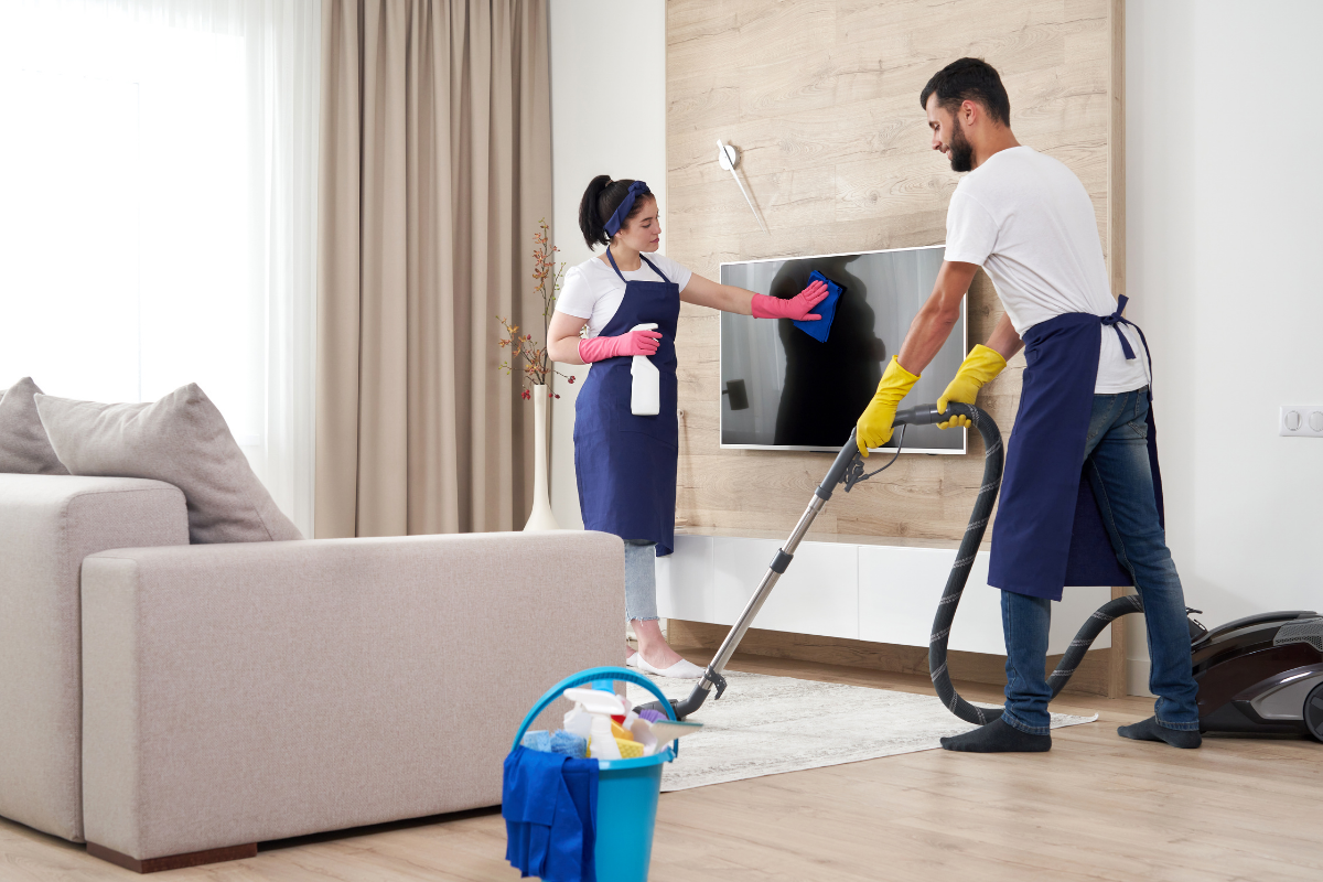 Professional cleaning team in matching aprons vacuuming a living room rug and wiping down a television screen.