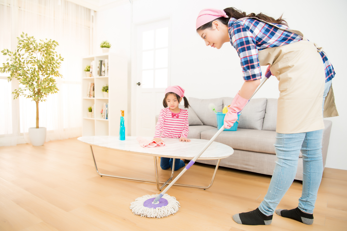 Mother mopping the floor while her young daughter wipes a table in a bright living room.
