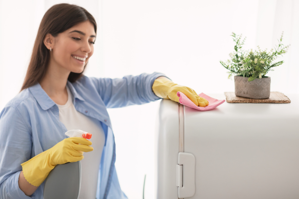 Smiling woman wearing yellow rubber gloves wiping down a refrigerator with a pink microfiber cloth and spray bottle.