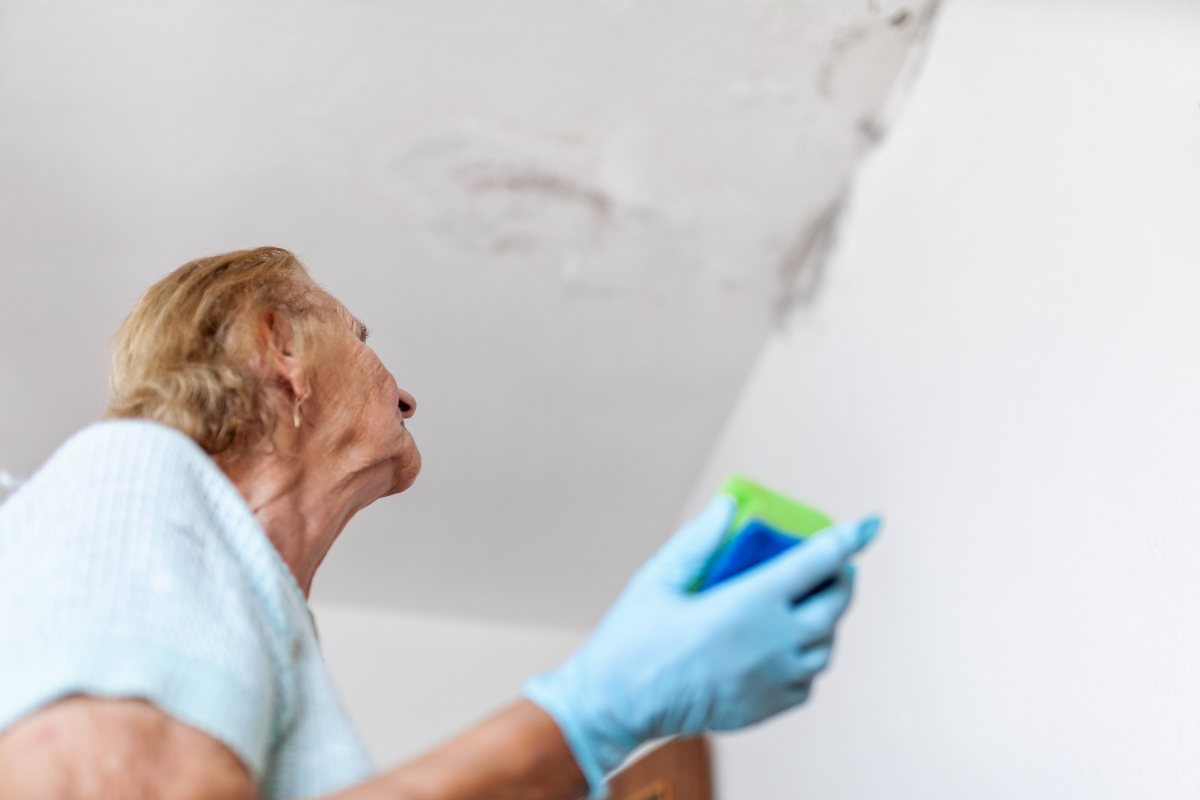 Senior woman wearing rubber gloves holding a sponge while inspecting water damage or mold on a ceiling.