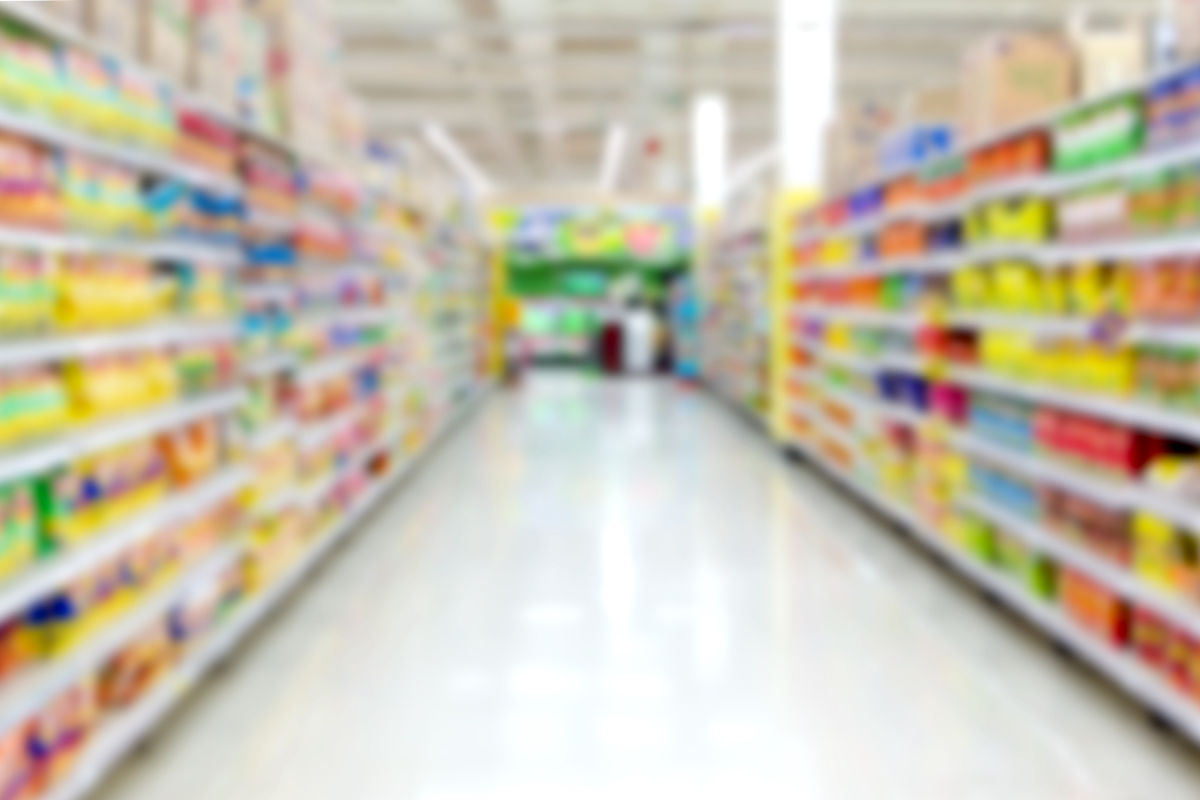Blurred view down a clean, brightly lit aisle in a retail store lined with stocked shelves on both sides.