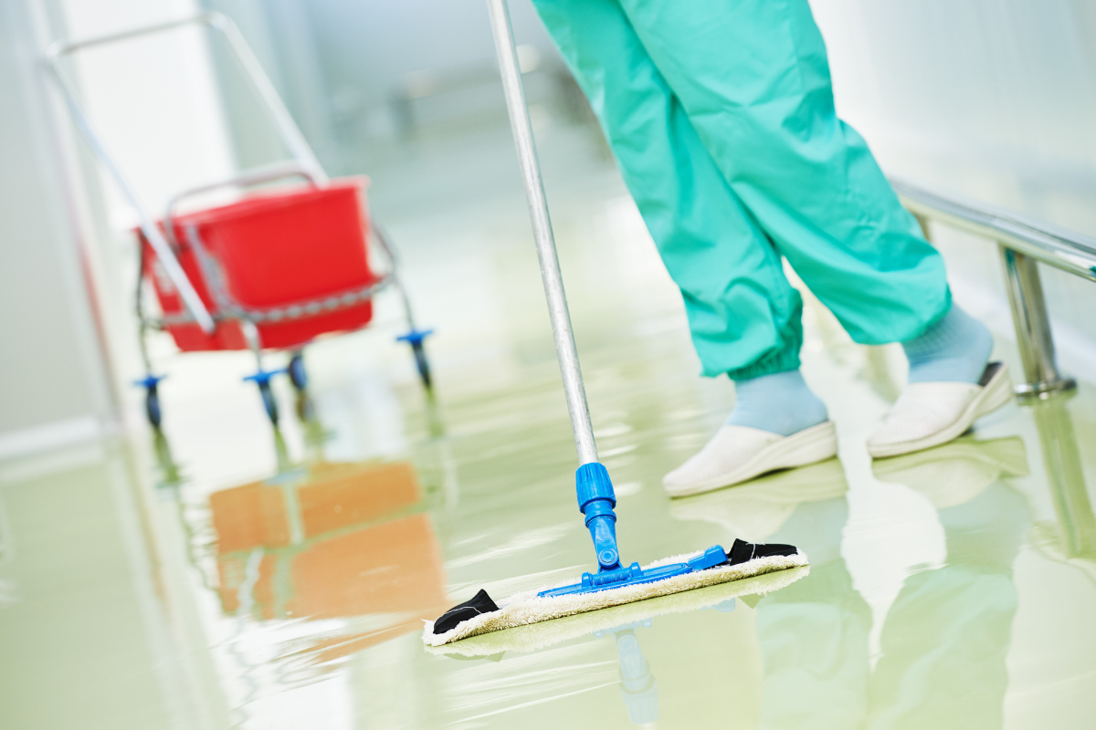 Professional cleaner in teal scrubs mopping a reflective floor next to a red cleaning cart.