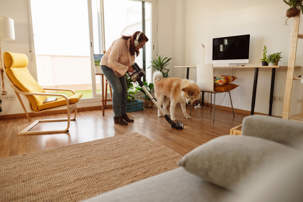 Woman wearing headphones vacuuming hardwood floor with a stick vacuum while a dog stands nearby in a modern living room.